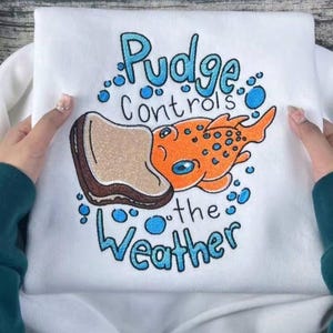 K&ouml;nnte beinhalten: Wei&szlig;es Sweatshirt mit farbenfroher Grafik. Das Design zeigt einen orangefarbenen Fisch mit blauen Punkten und den Text "Pudge Controls the Weather" in Blau. Ein Brotst&uuml;ck ist ebenfalls im Design enthalten.