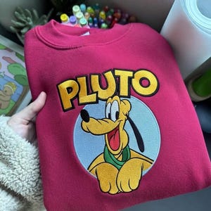 Op de afbeelding: Een felroze sweater met ronde hals en een grote geborduurde afbeelding van het stripfiguur Pluto. Het ontwerp bevat de naam PLUTO in gele blokletters boven het personage, geplaatst tegen een lichtblauwe cirkelvormige achtergrond.