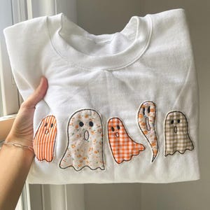 May include: A white crewneck sweatshirt featuring five ghost-shaped fabric appliqués stitched across the front. The ghosts are made from various patterned fabrics, including orange stripes, floral prints, orange gingham, and a tan checkered pattern.