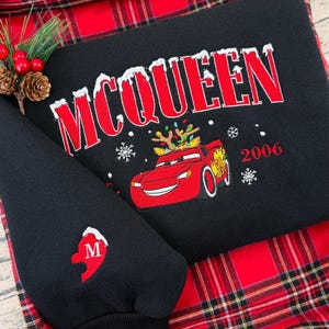 Puede incluir: Sudadera negra con el nombre "McQUEEN" en rojo con dise&ntilde;o nevado. Un coche de dibujos animados rojo con cuernos y luces navide&ntilde;as bordado debajo. El a&ntilde;o "2006" tambi&eacute;n est&aacute; en rojo. La manga tiene un coraz&oacute;n rojo con la letra "M".