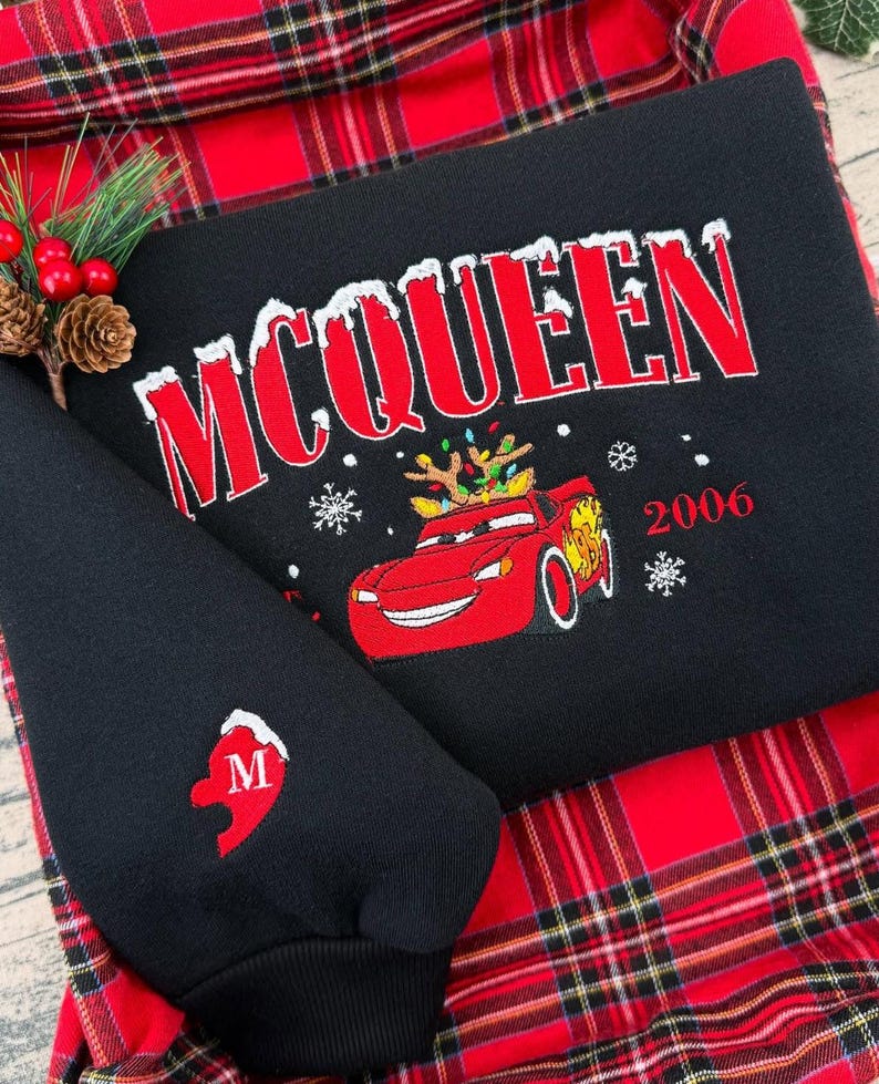Puede incluir: Sudadera negra con el nombre "McQUEEN" en rojo con dise&ntilde;o nevado. Un coche de dibujos animados rojo con cuernos y luces navide&ntilde;as bordado debajo. El a&ntilde;o "2006" tambi&eacute;n est&aacute; en rojo. La manga tiene un coraz&oacute;n rojo con la letra "M".