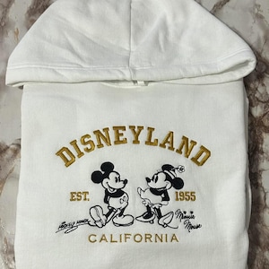 Op de afbeelding: Witte hoodie met gouden borduursel van "DISNEYLAND" en illustraties van Mickey en Minnie Mouse. Het ontwerp bevat "EST. 1955 CALIFORNIA" in goud en zwart, een klassiek thema.