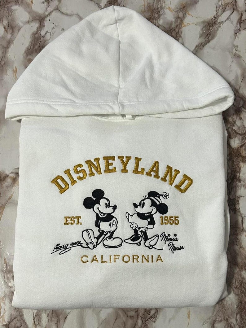 Op de afbeelding: Witte hoodie met gouden borduursel van "DISNEYLAND" en illustraties van Mickey en Minnie Mouse. Het ontwerp bevat "EST. 1955 CALIFORNIA" in goud en zwart, een klassiek thema.