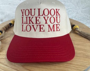 May include: A baseball cap with a cream-colored crown and a red brim. The cap features the embroidered text "YOU LOOK LIKE YOU LOVE ME" in red. The cap has a red button on top.