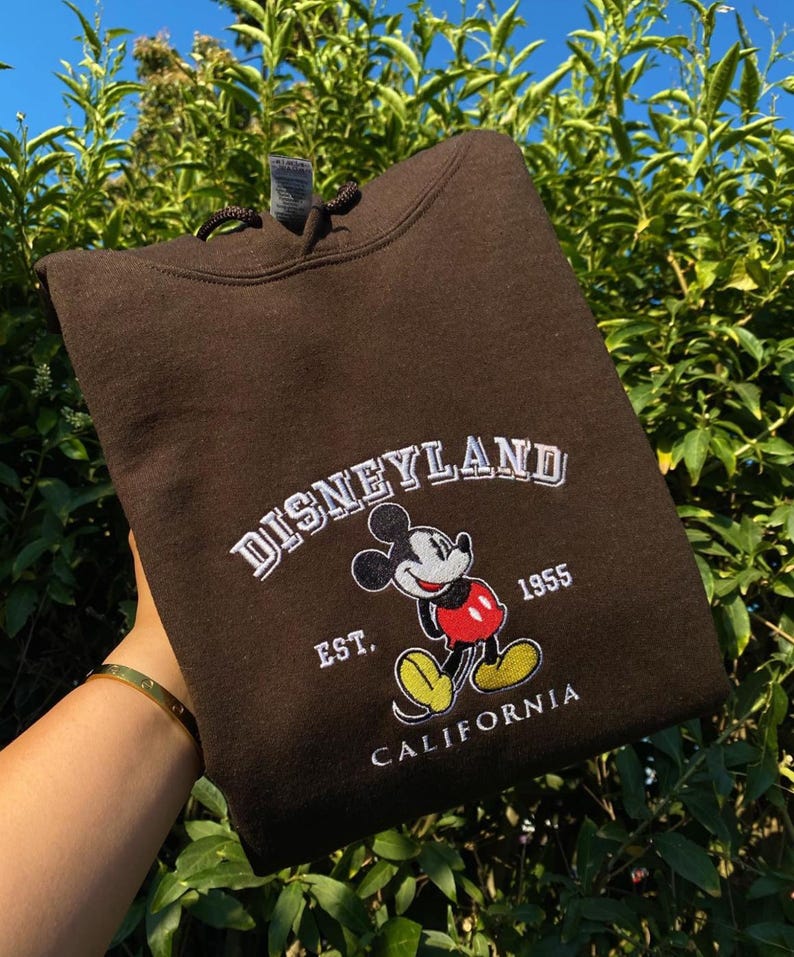 May include: A brown Disneyland hoodie with white embroidered text and a Mickey Mouse graphic. The text reads "DISNEYLAND", "EST. 1955", and "CALIFORNIA". The hoodie has a drawstring and is folded.