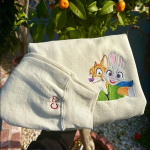 May include: Cream sweatshirt featuring embroidered cartoon characters of a fox and a rabbit. The sleeve cuff has a small red heart and the letters "CO". The characters are wearing green and blue clothing.