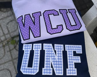 Puede incluir: Una sudadera de color morado claro con las letras WCU en morado con lunares blancos y borde negro, sobre una sudadera azul marino con las letras UNF en un patrón de cuadros vichy azul claro y blanco con borde blanco.