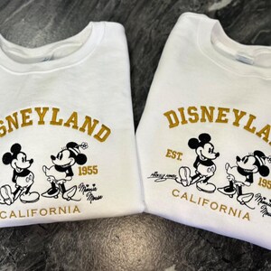 Op de afbeelding: Twee witte sweatshirts met het Disneyland-logo in gouden letters, met Mickey en Minnie Mouse. De sweatshirts hebben de tekst "EST. 1955 CALIFORNIA" en handtekeningen. Een klassiek Disney-thema.