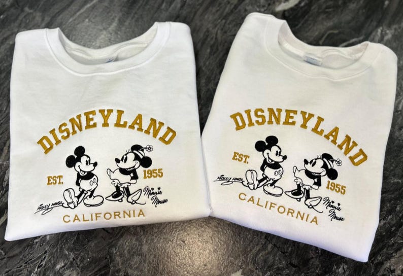 Op de afbeelding: Twee witte sweatshirts met het Disneyland-logo in gouden letters, met Mickey en Minnie Mouse. De sweatshirts hebben de tekst "EST. 1955 CALIFORNIA" en handtekeningen. Een klassiek Disney-thema.