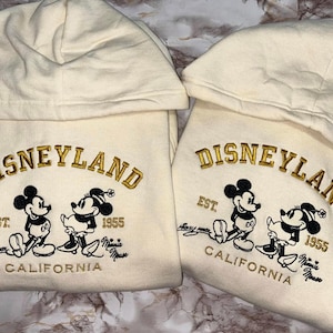 Op de afbeelding: Twee cr&egrave;mekleurige Disneyland-hoodies met geborduurde gouden letters en Mickey en Minnie Mouse-afbeeldingen. De hoodies hebben de tekst "Disneyland", "Est. 1955" en "California".