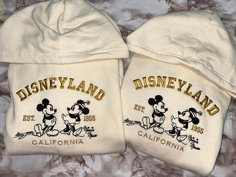 Op de afbeelding: Twee cr&egrave;mekleurige Disneyland-hoodies met geborduurde gouden letters en Mickey en Minnie Mouse-afbeeldingen. De hoodies hebben de tekst "Disneyland", "Est. 1955" en "California".