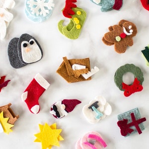 May include: A set of felt Christmas ornaments in various shapes and colors, including a penguin, a snowman, a gingerbread man, a Christmas tree, a stocking, a candy cane, a wreath, a star, a bell, a cardinal, a snowflake, a Santa hat, and a present.