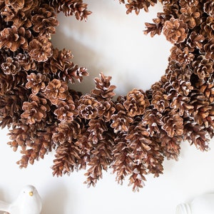 May include: A brown pine cone wreath with a white frosted finish. The wreath is made of natural pine cones and is perfect for a winter or holiday decor.