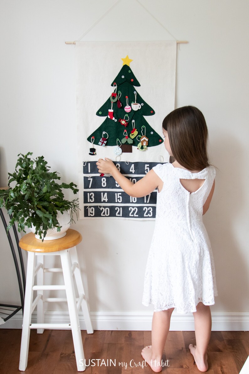 May include: A felt Christmas tree advent calendar with 23 numbered pockets and a felt star on top. A child is opening a pocket on the calendar.