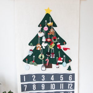 May include: A felt Christmas tree advent calendar with 24 numbered pockets. Each pocket features a different felt ornament, including a gingerbread man, a stocking, a snowman, a penguin, and a candy cane. The calendar is hung on a white wall.