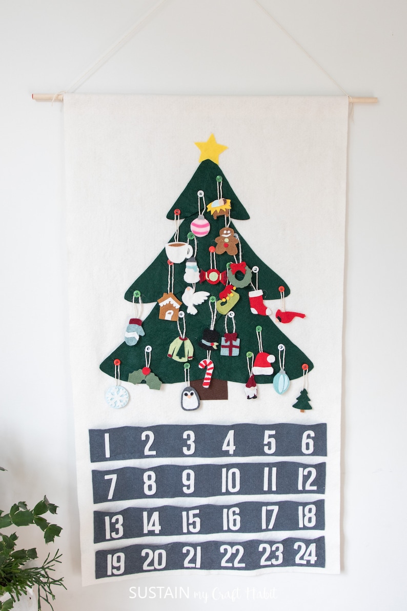 May include: A felt Christmas tree advent calendar with 24 numbered pockets. Each pocket features a different felt ornament, including a gingerbread man, a stocking, a snowman, a penguin, and a candy cane. The calendar is hung on a white wall.
