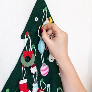May include: A felt Christmas tree with a yellow star on top and various felt ornaments hanging from it. The ornaments include a wreath, a stocking, a snowman, a bird, a gingerbread house, and a few ornaments. A hand is shown hanging a blue ornament on the tree.