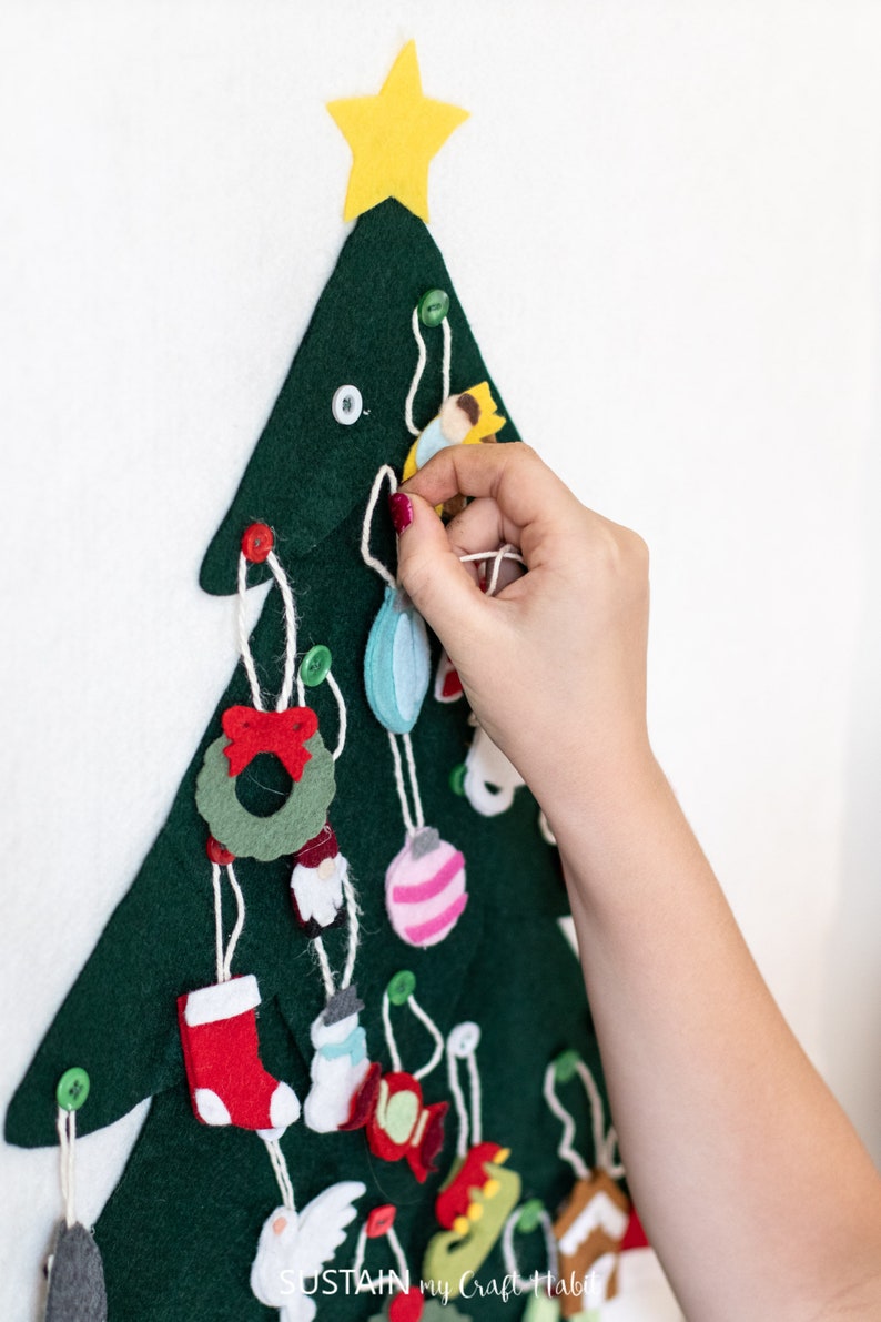 May include: A felt Christmas tree with a yellow star on top and various felt ornaments hanging from it. The ornaments include a wreath, a stocking, a snowman, a bird, a gingerbread house, and a few ornaments. A hand is shown hanging a blue ornament on the tree.