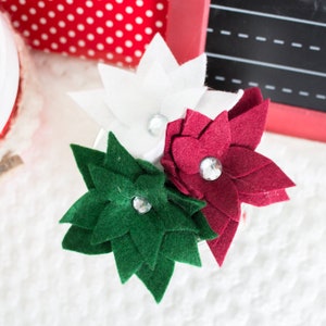 以下が含まれることがあります： 赤、白、緑のフェルトでできたポインセチアの花が3つ、ラインストーンのセンターが付いています。花は束になって配置されています。