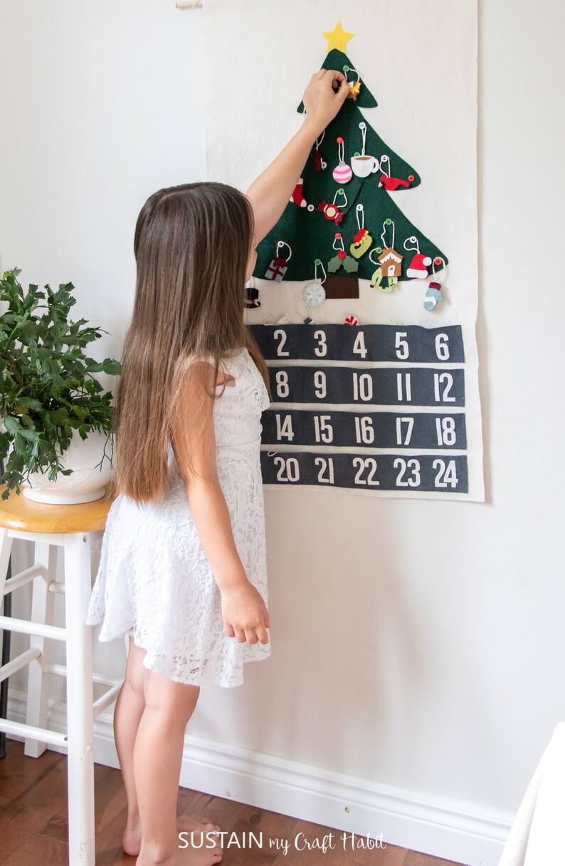 May include: A felt Christmas tree advent calendar with numbers 1 through 24. A young girl is hanging a felt ornament on the tree.