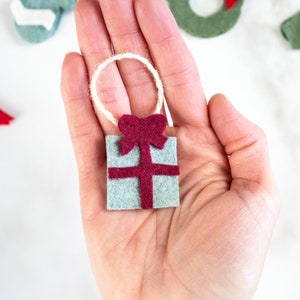 May include: A small felt ornament shaped like a gift box with a red bow. The ornament is light blue with a red ribbon and bow. The ornament has a loop of twine for hanging.