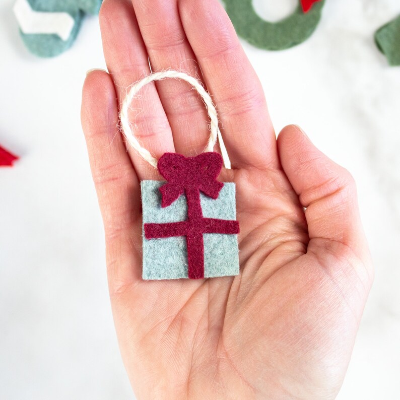 May include: A small felt ornament shaped like a gift box with a red bow. The ornament is light blue with a red ribbon and bow. The ornament has a loop of twine for hanging.