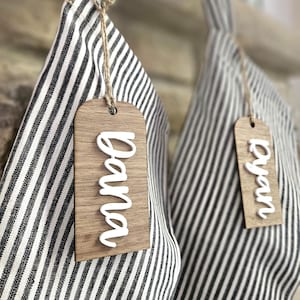 May include: Two striped fabric items with wooden tags. One tag says "Nana" in white script, the other says "Ryan". The tags are attached with twine. The fabric has black and white stripes.