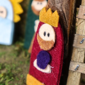 May include: Felt Christmas ornament of a king with a red robe, gold crown, and a purple jewel. The king has a brown beard and a white face with black eyes.