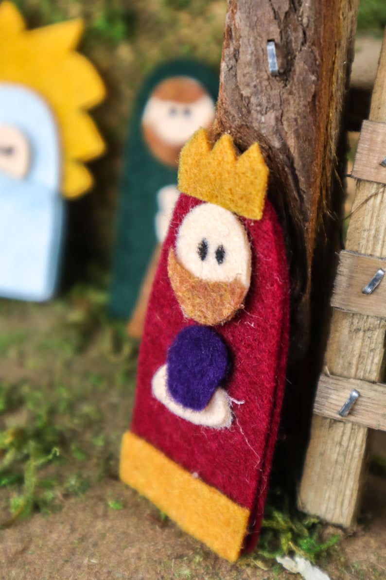 May include: Felt Christmas ornament of a king with a red robe, gold crown, and a purple jewel. The king has a brown beard and a white face with black eyes.
