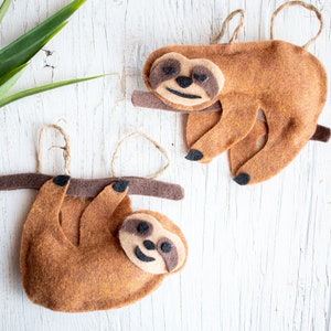 May include: Two felt sloth ornaments in shades of brown, hanging from twine loops. The sloths are perched on brown branches, with black claws and tan faces. The background is a white, textured surface.