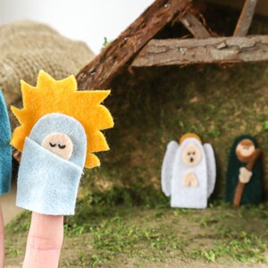May include: Felt finger puppets depicting the Nativity scene. The puppets include Mary, Joseph, baby Jesus, an angel, a shepherd, and a wise man. The puppets are in front of a wooden stable with a gold star on top.