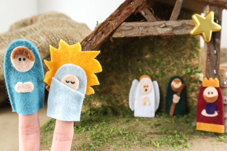 May include: Felt finger puppets depicting the Nativity scene. The puppets include Mary, Joseph, baby Jesus, an angel, a shepherd, and a wise man. The puppets are in front of a wooden stable with a gold star on top.