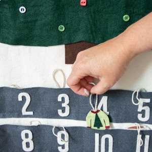 May include: A felt Christmas advent calendar with numbers 2-17 on gray felt strips. Each number has a loop of twine attached. A hand is pulling on the twine attached to number 4, revealing a felt ornament shaped like a green sweater with a small green tree on it.
