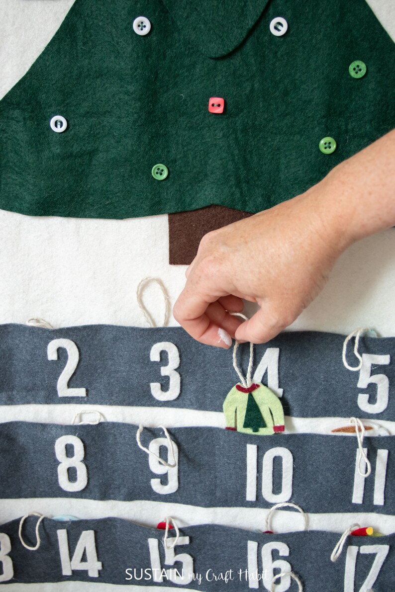 May include: A felt Christmas advent calendar with numbers 2-17 on gray felt strips. Each number has a loop of twine attached. A hand is pulling on the twine attached to number 4, revealing a felt ornament shaped like a green sweater with a small green tree on it.