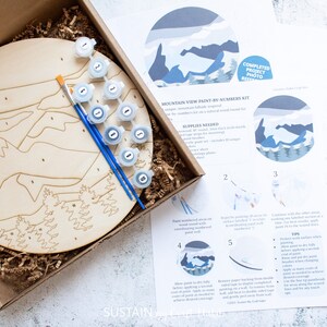 Peut inclure: Un kit de peinture par num&eacute;ros repr&eacute;sentant un paysage de montagne. Le kit comprend une toile ronde en bois, des pots de peinture et des pinceaux. Les instructions sont imprim&eacute;es sur une feuille de papier s&eacute;par&eacute;e.