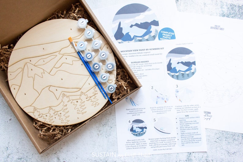 Peut inclure: Un kit de peinture par num&eacute;ros repr&eacute;sentant un paysage de montagne. Le kit comprend une toile ronde en bois, des pots de peinture et des pinceaux. Les instructions sont imprim&eacute;es sur une feuille de papier s&eacute;par&eacute;e.