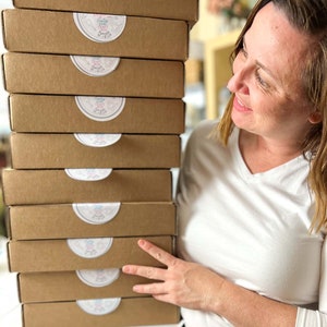 Peut inclure: Une pile de neuf bo&icirc;tes en carton brun avec des &eacute;tiquettes circulaires blanches qui disent "Craft Happy". Les bo&icirc;tes sont empil&eacute;es sur une table et une personne se tient derri&egrave;re elles.