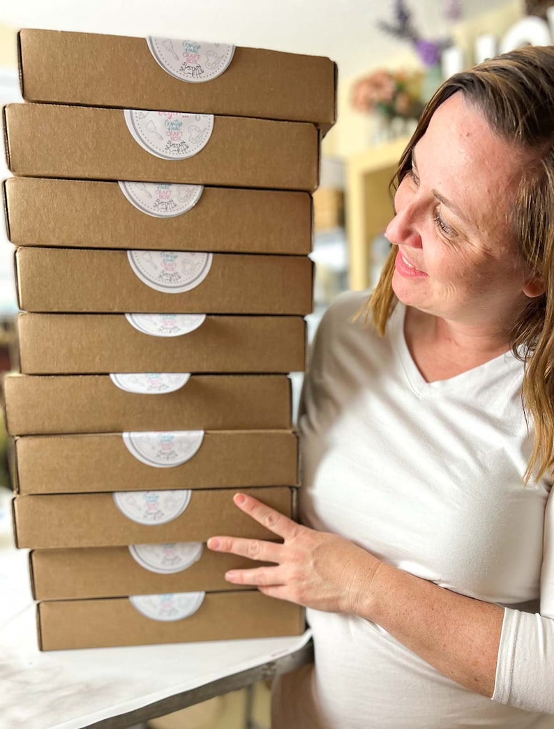 Peut inclure: Une pile de neuf bo&icirc;tes en carton brun avec des &eacute;tiquettes circulaires blanches qui disent "Craft Happy". Les bo&icirc;tes sont empil&eacute;es sur une table et une personne se tient derri&egrave;re elles.