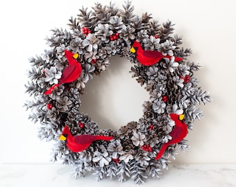 May include: A circular wreath constructed from numerous pinecones with a white-washed finish. The wreath is decorated with small clusters of bright red berries and four red felt cardinal birds, each featuring a yellow beak and black face detail.