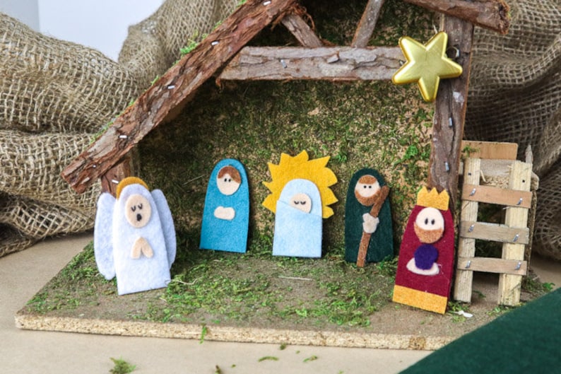 May include: A felt nativity scene with a manger, a star, a ladder, and six figures: an angel, Mary, Joseph, a shepherd, a king, and baby Jesus. The figures are all standing on a green moss-covered surface.
