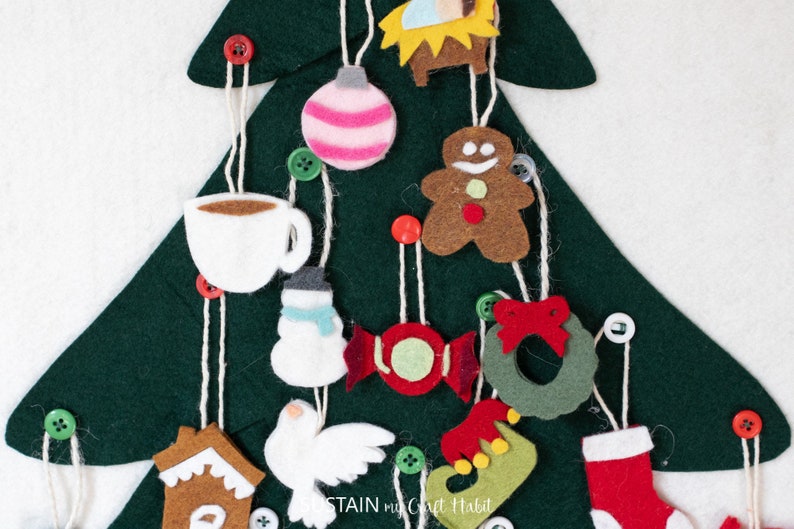 May include: A felt Christmas tree with various felt ornaments hanging from it. The ornaments include a gingerbread man, a candy cane, a snowman, a dove, a stocking, a coffee cup, a wreath, a star, a gingerbread house, and a nativity scene.