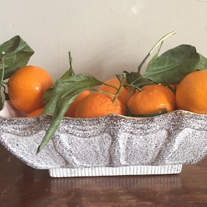 May include: A white ceramic bowl with a textured surface, filled with orange mandarins and green leaves.