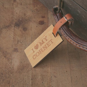 May include: A wooden luggage tag with a brown leather strap. The tag has the text "I ♥ MY CORNET" engraved on it.