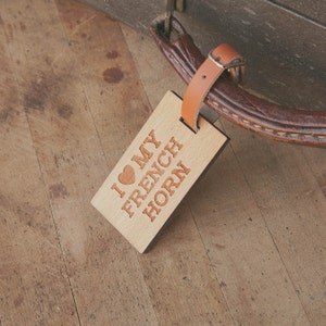 May include: A wooden luggage tag with a brown leather strap. The tag has the text "I love my French Horn" engraved on it with a red heart.