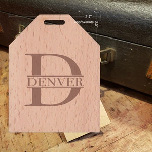 May include: A light brown wooden luggage tag with a cut-out handle. The tag has a large letter "D" with the word "DENVER" centered below the letter. The tag is approximately 2.7 inches wide and 3.5 inches tall.