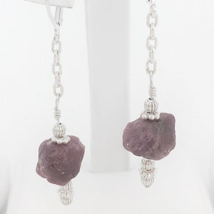 Puede incluir: Un par de pendientes de plata con un colgante de piedra preciosa púrpura. Los pendientes tienen una cadena de plata y una pequeña perla de plata en la parte superior de la piedra preciosa.