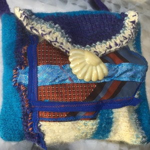 May include: A small blue and white knitted bag with a crocheted trim and a shell-shaped button. The bag is decorated with a blue and brown patterned fabric.