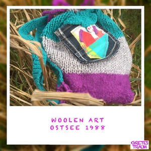 Op de afbeelding: Een turquoise, grijs en paarse gebreide tas met een denim zak en een kleurrijke patch. De tas heeft een lange band en ligt op een bed van droog gras. De tekst "WOOLEN ART OSTSEE 1988" is in roze op een witte achtergrond geschreven.