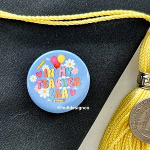 May include: A blue button with the text "In My Teacher Era" in colorful letters with hearts, flowers, and a pencil. The button is on a black surface with a yellow tassel.