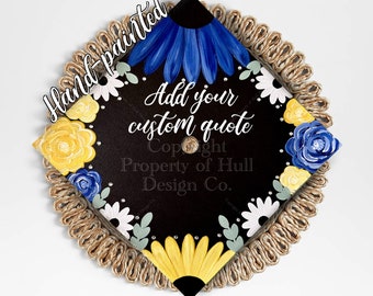 May include: Hand-painted graduation cap with a black background, yellow, blue and white flowers, and rhinestones. The text "Add your custom quote" is written in white on the cap. The text "Copyright Property of Hull Design Co." is written in white on the cap.
